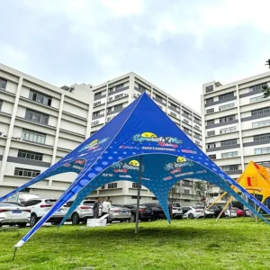 Large blue printed star-shaped tent set on grass with buildings, parked vehicles and other tents in the background