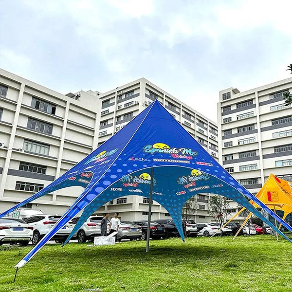 Large blue printed star-shaped tent set on grass with buildings, parked vehicles and other tents in the background