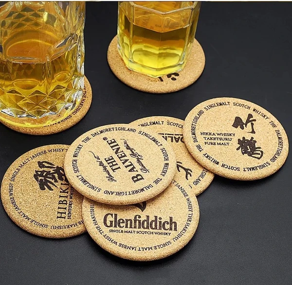 Round cork coasters with whisky brand artwork arranged beside two glasses of amber drink on a dark surface